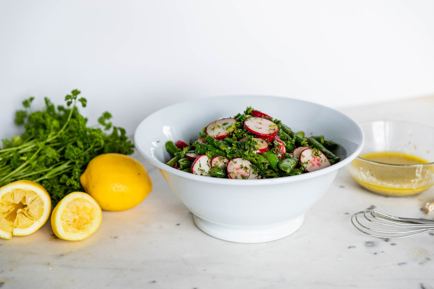 Large white stoneware bowl filled with asparagus salad with radish and sugar peas topped with fresh dill and homemade vinaigrette with extra lemons, parsley and vinaigrette to the sides on a marble surface