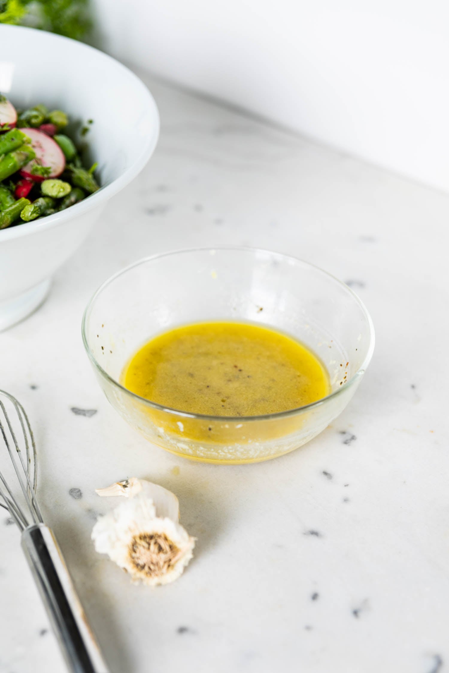 Glass bowl full of yellow homemade vinaigrette with white bowl filled with asparagus salad with radish and sugar peas in background with a garlic clove and whisk in front all on a marble surface