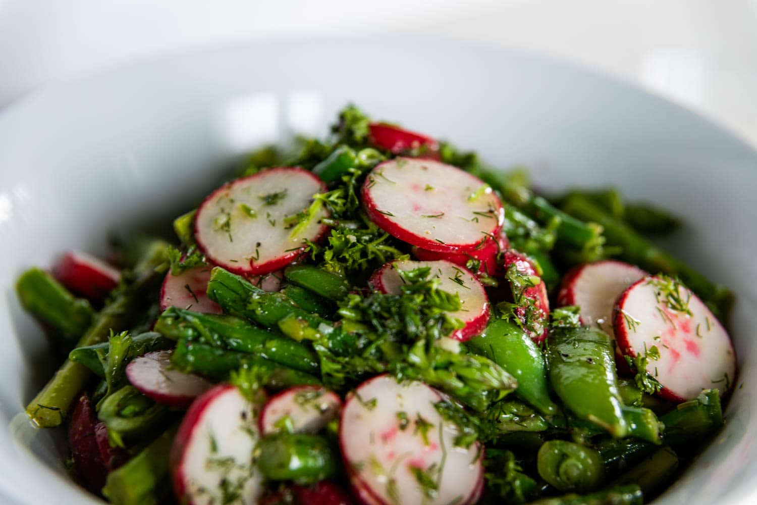 Close view of vibrant an asparagus salad with radish and sugar peas in a large white bowl sprinkled with fresh dill and a homemade vinaigrette