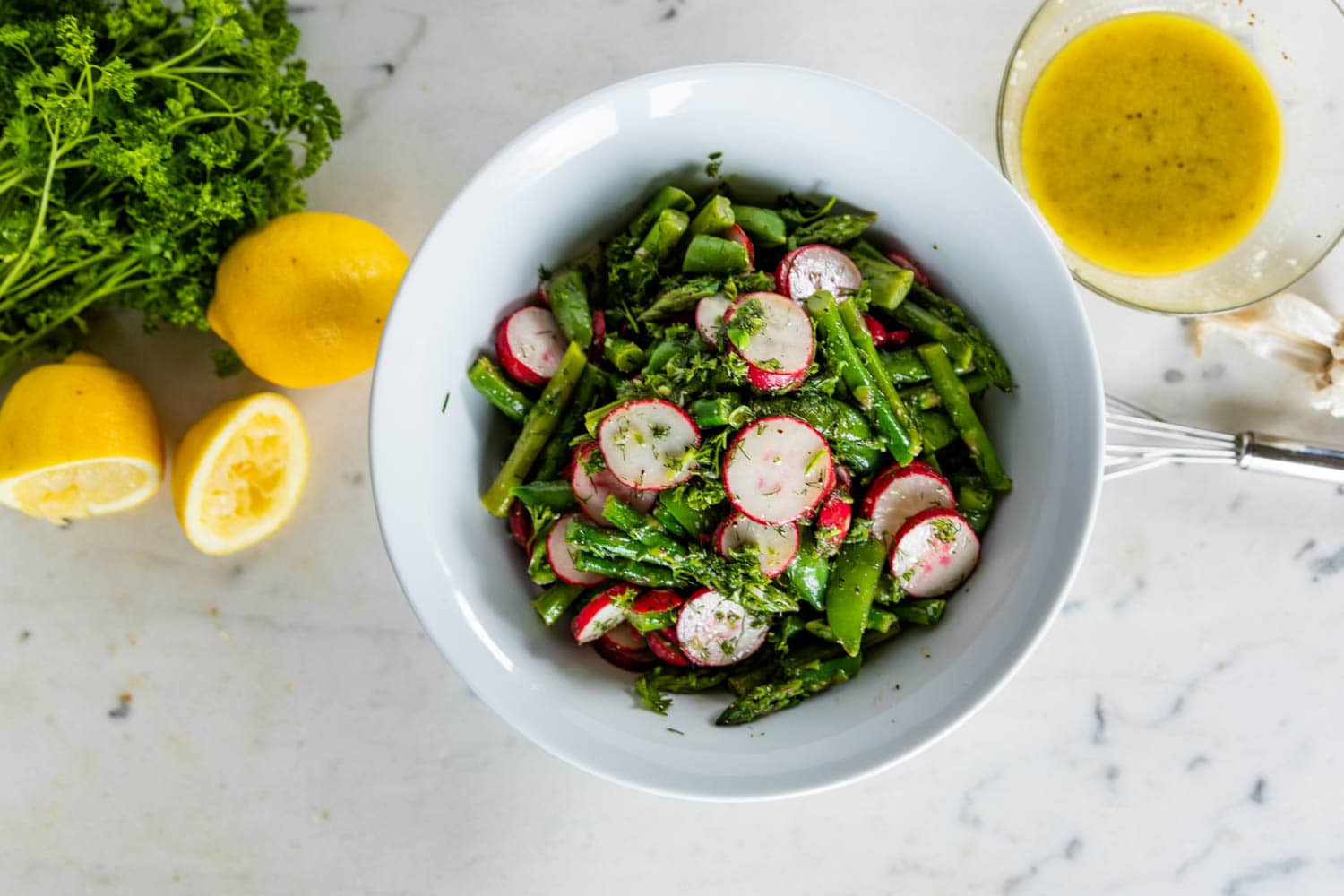 Top down view of asparagus salad with radish and sugar peas in a large white bowl with extra lemons, parsley, garlic and homemade vinaigrette on a marble surface
