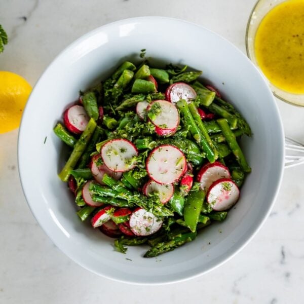 Top down view of asparagus salad with radish and sugar peas in a large white bowl with extra lemons, parsley, garlic and homemade vinaigrette on a marble surface