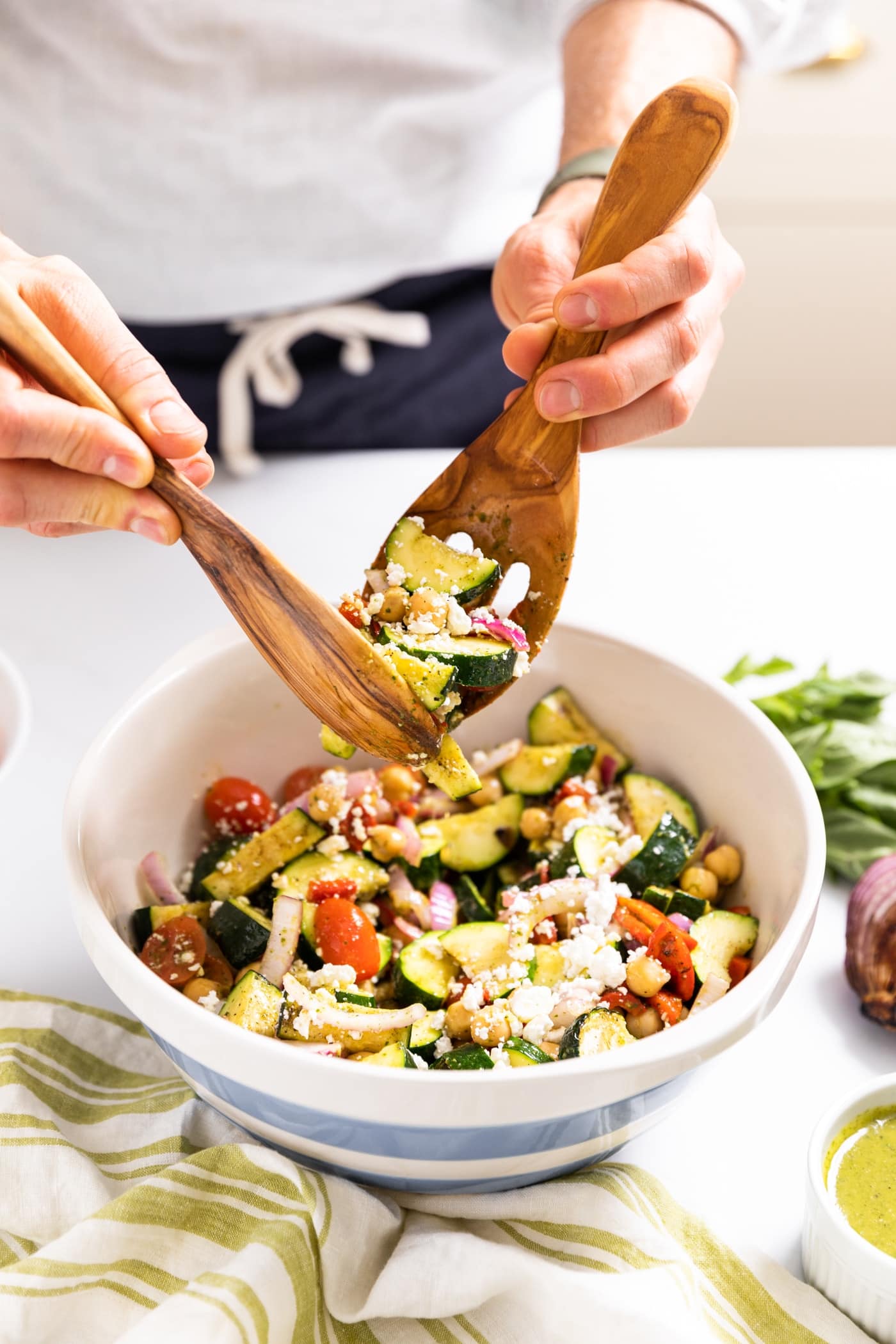 Two hands holding wooden tongs scooping up zucchini salad from white and blue bowl sitting on white countertop