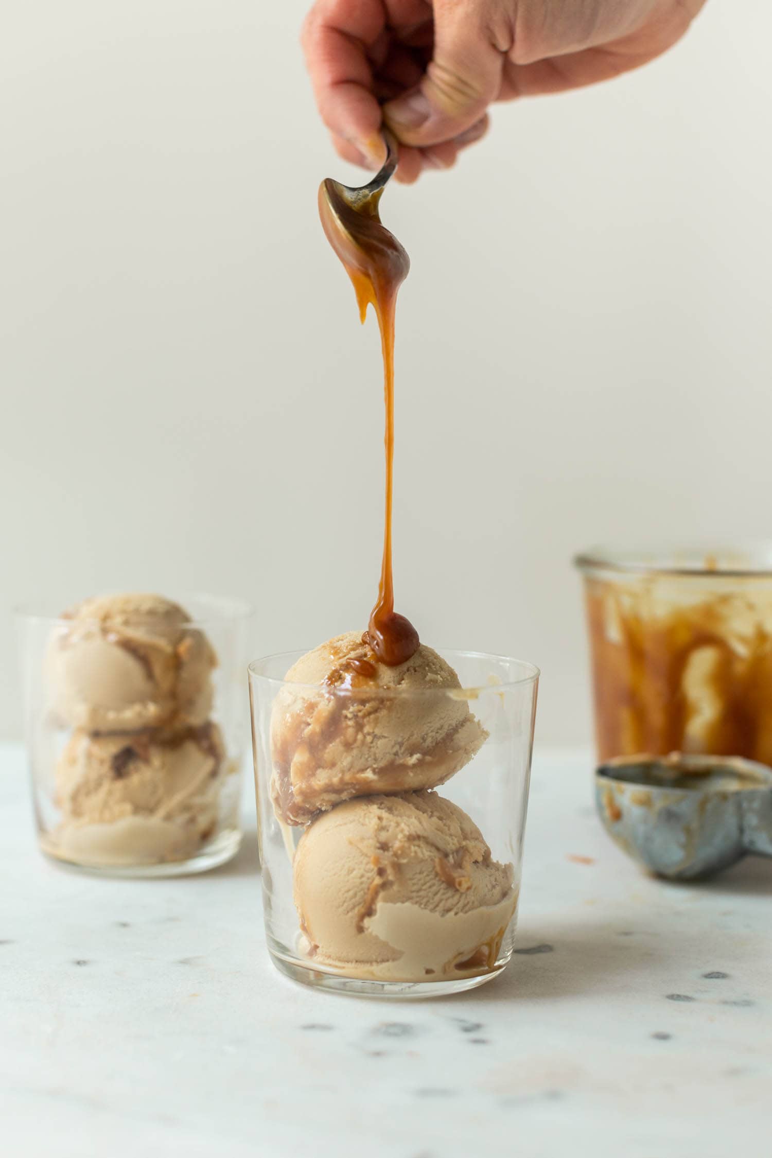 Caramel macchiato ice cream with caramel sauce being drizzled on top in a clear glass with a second glass full of ice cream in background as well as extra homemade caramel sauce in a jar and an ice cream scoop on a marble surface