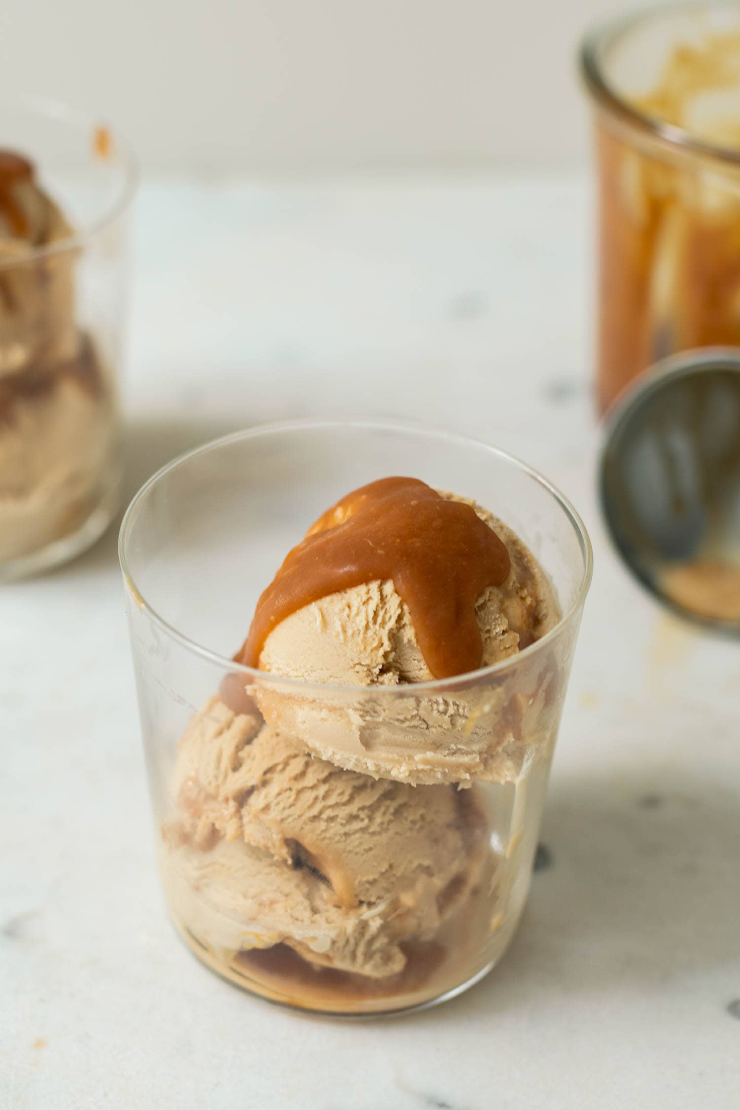 Caramel macchiato ice cream with caramel sauce drizzled on top in a clear glass with a second glass full of ice cream in background as well as extra homemade caramel sauce in a jar and an ice cream scoop on a marble surface