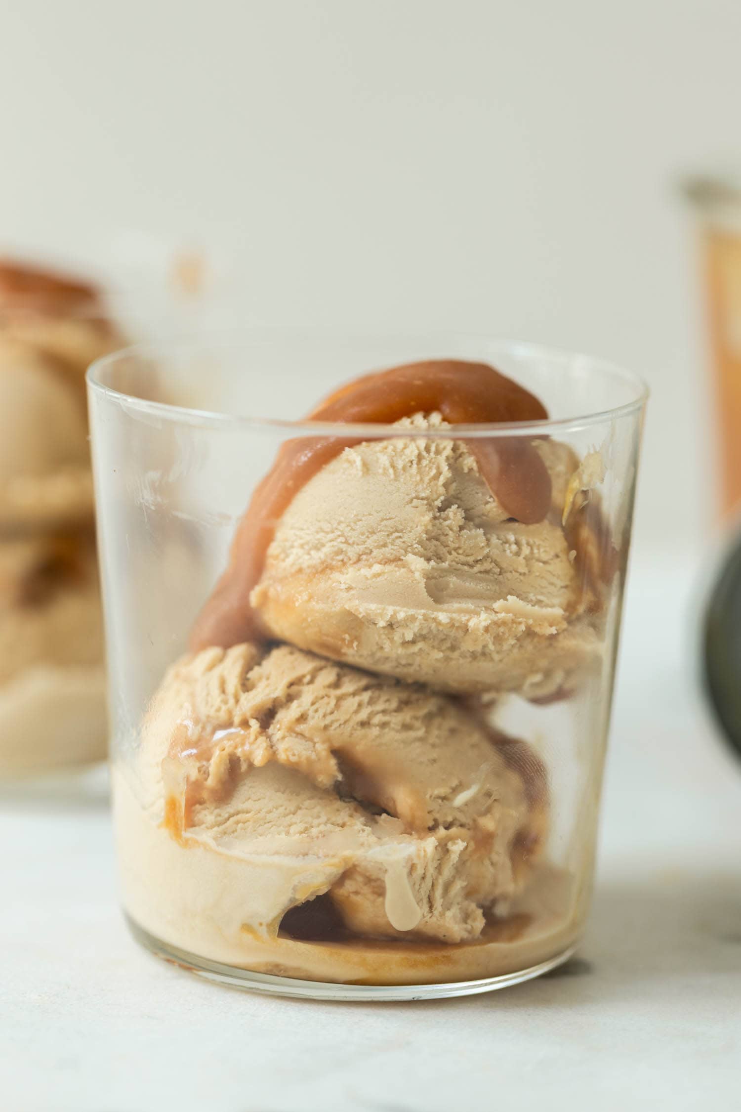 Caramel macchiato ice cream with caramel sauce drizzled on top in a clear glass with a second glass full of ice cream in background as well as extra homemade caramel sauce in a jar and an ice cream scoop on a marble surface
