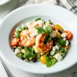 Grilled tequila lime shrimp on a bed of cherry tomatoes, corn and cilantro and sprinkled with feta cheese with second plate, fork and linen towel in background on a marble surface