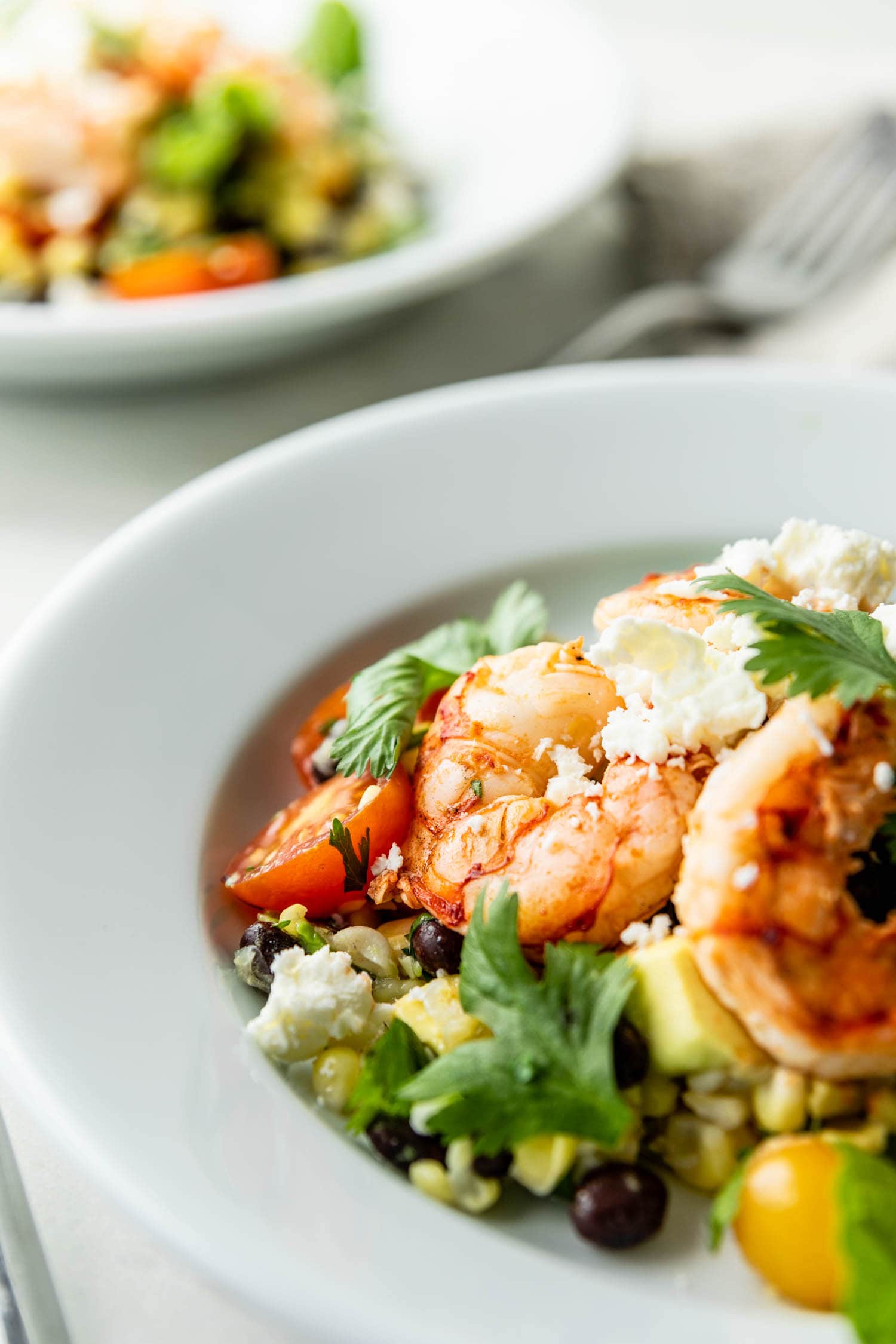 Grilled tequila lime shrimp on a bed of cherry tomatoes, corn and cilantro and sprinkled with feta cheese with second plate in background on a marble surface with a linen towel