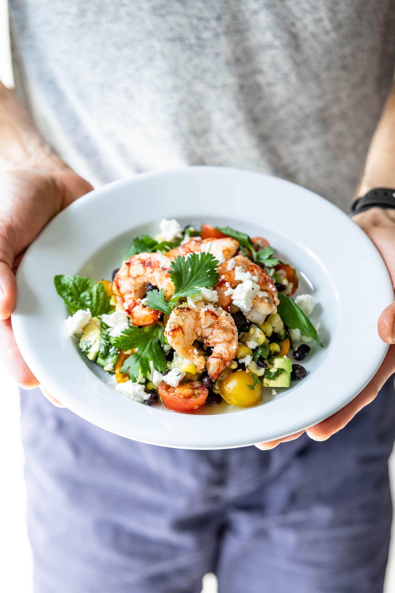 Two hands holding a plate of grilled tequila lime shrimp on a bed of cherry tomatoes, corn and cilantro and sprinkled with feta cheese