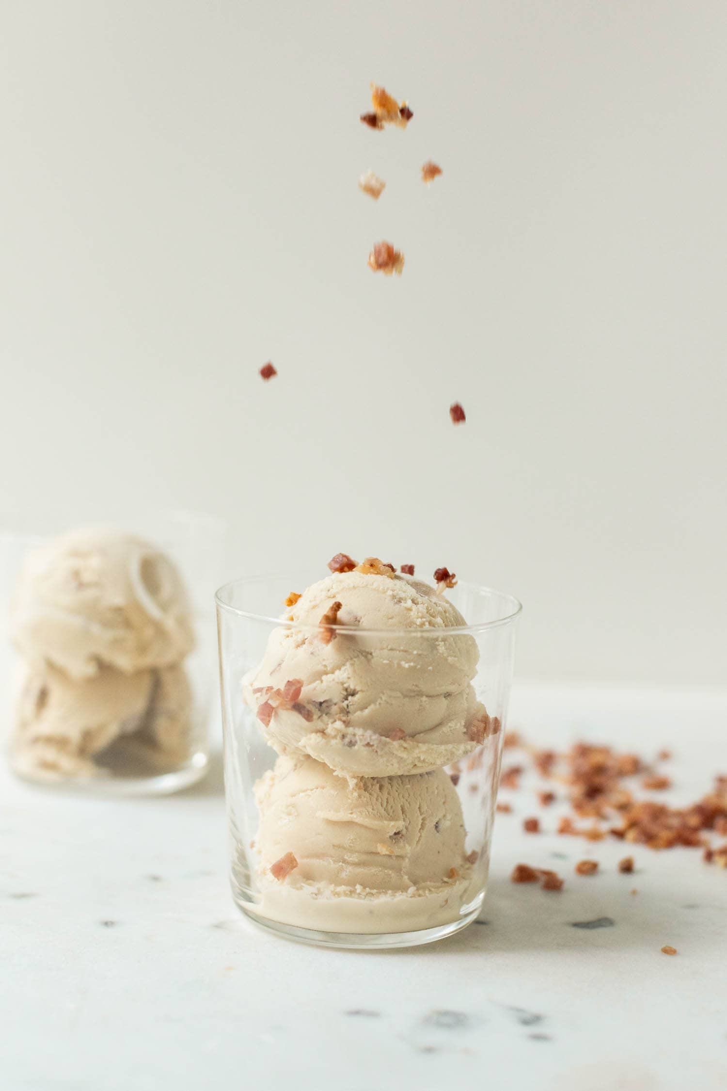 Maple bacon ice cream with bacon pieces being sprinkled on top with second glass full of ice cream and bacon pieces in background on a marble surface