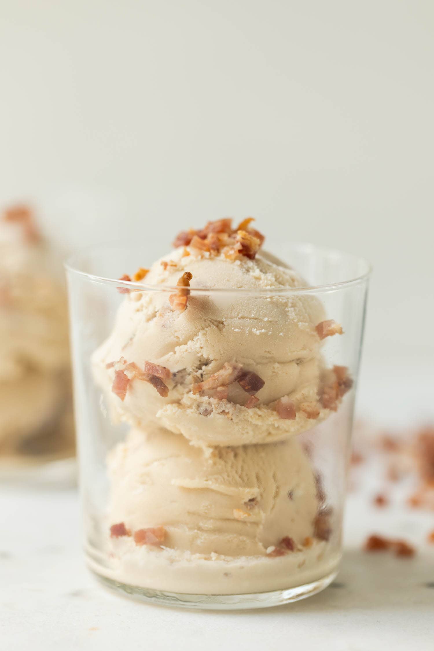 Maple bacon ice cream with bacon pieces on top and down the sides in a clear glass with a second glass in background on a marble surface