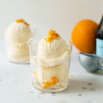 Mimosa ice cream speckled with orange zest pieces in a clear glass with second ice cream cup, orange, ice cream scoop and champagne bottle in background on a white marble surface