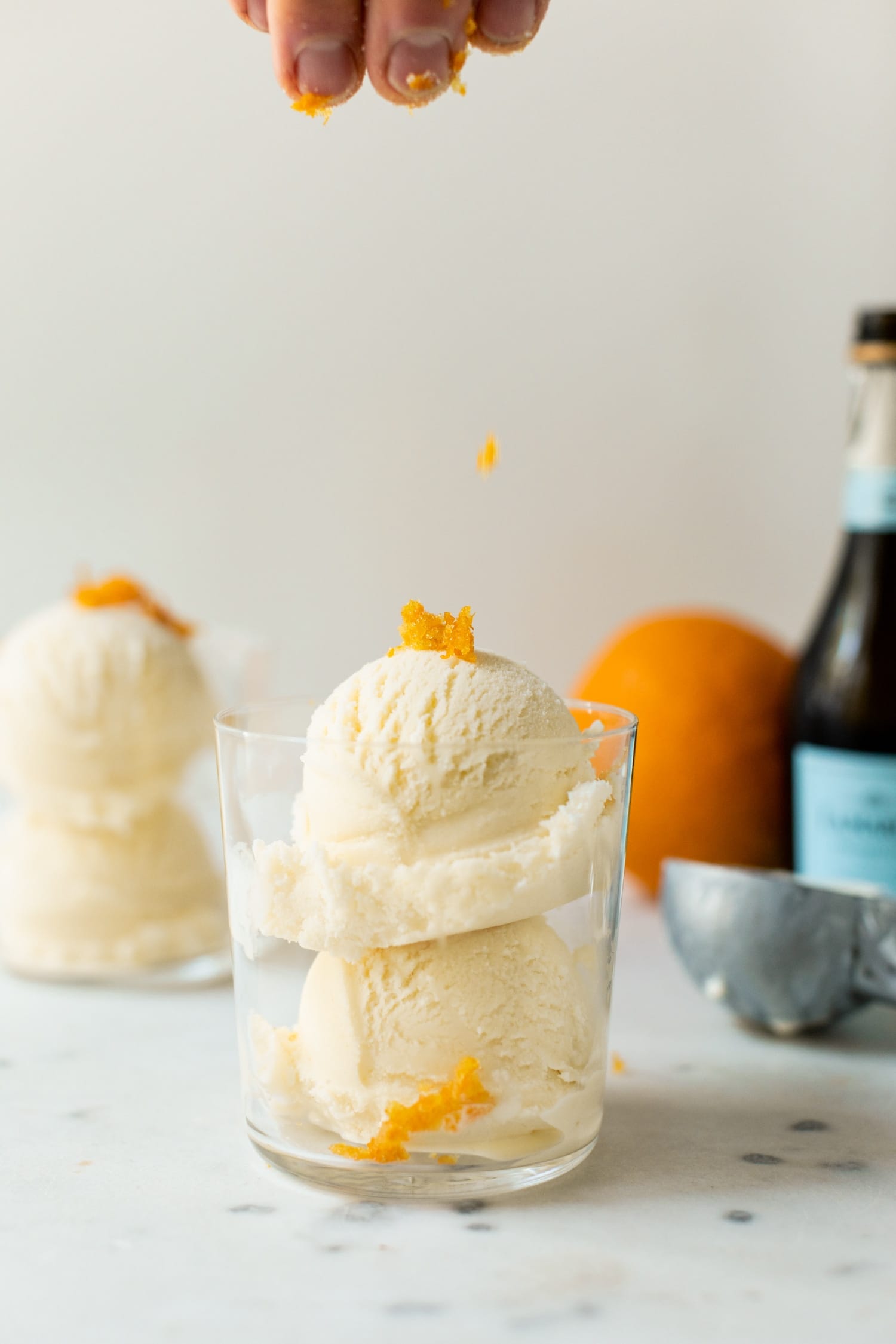 Mimosa ice cream being speckled with orange zest pieces from the top of the picture in a clear glass with second ice cream cup, orange, ice cream scoop and champagne bottle in background on a white marble surface