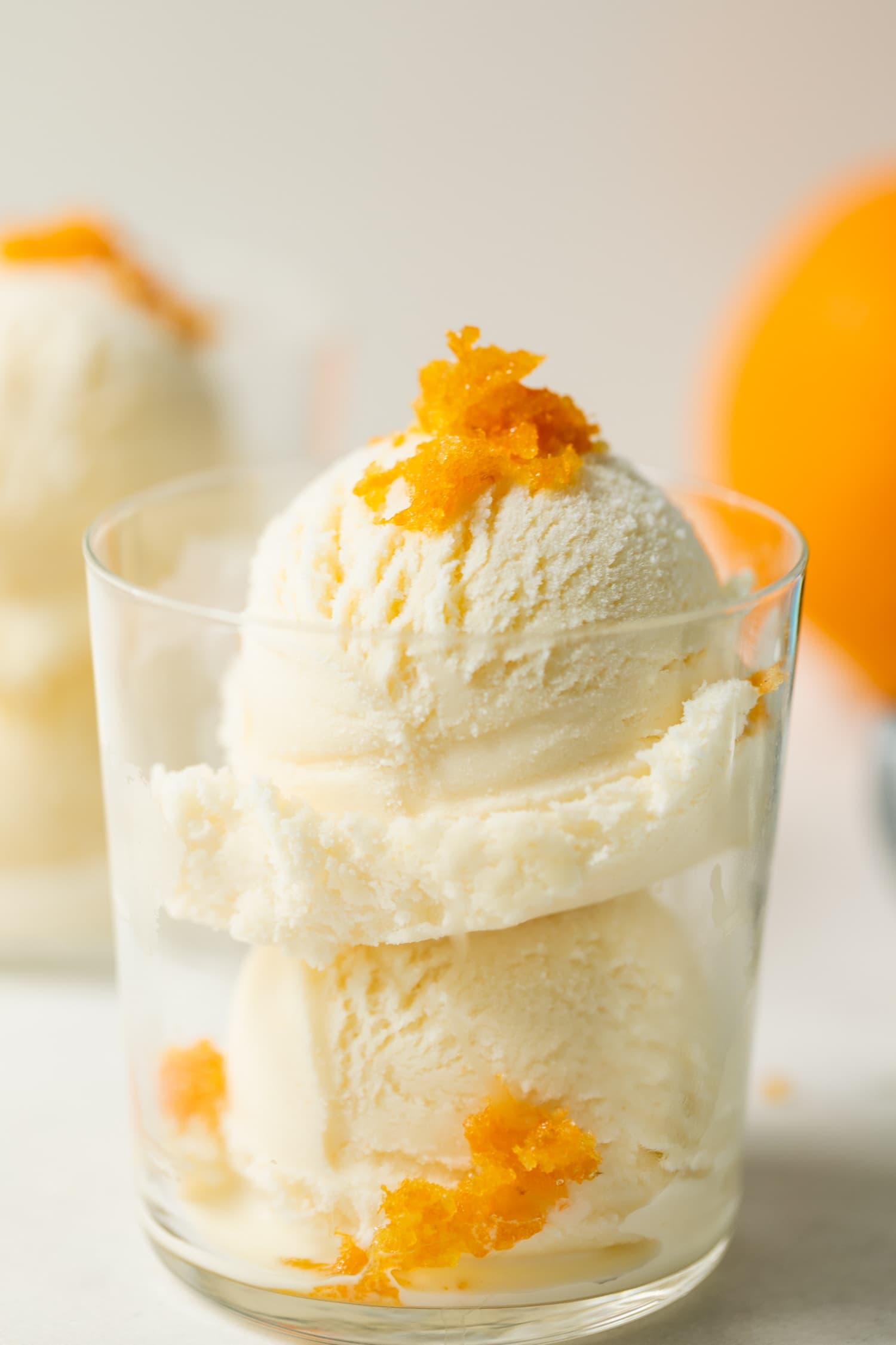 Mimosa ice cream speckled with orange zest pieces in a clear glass on marble surface with second ice cream glass and orange in background