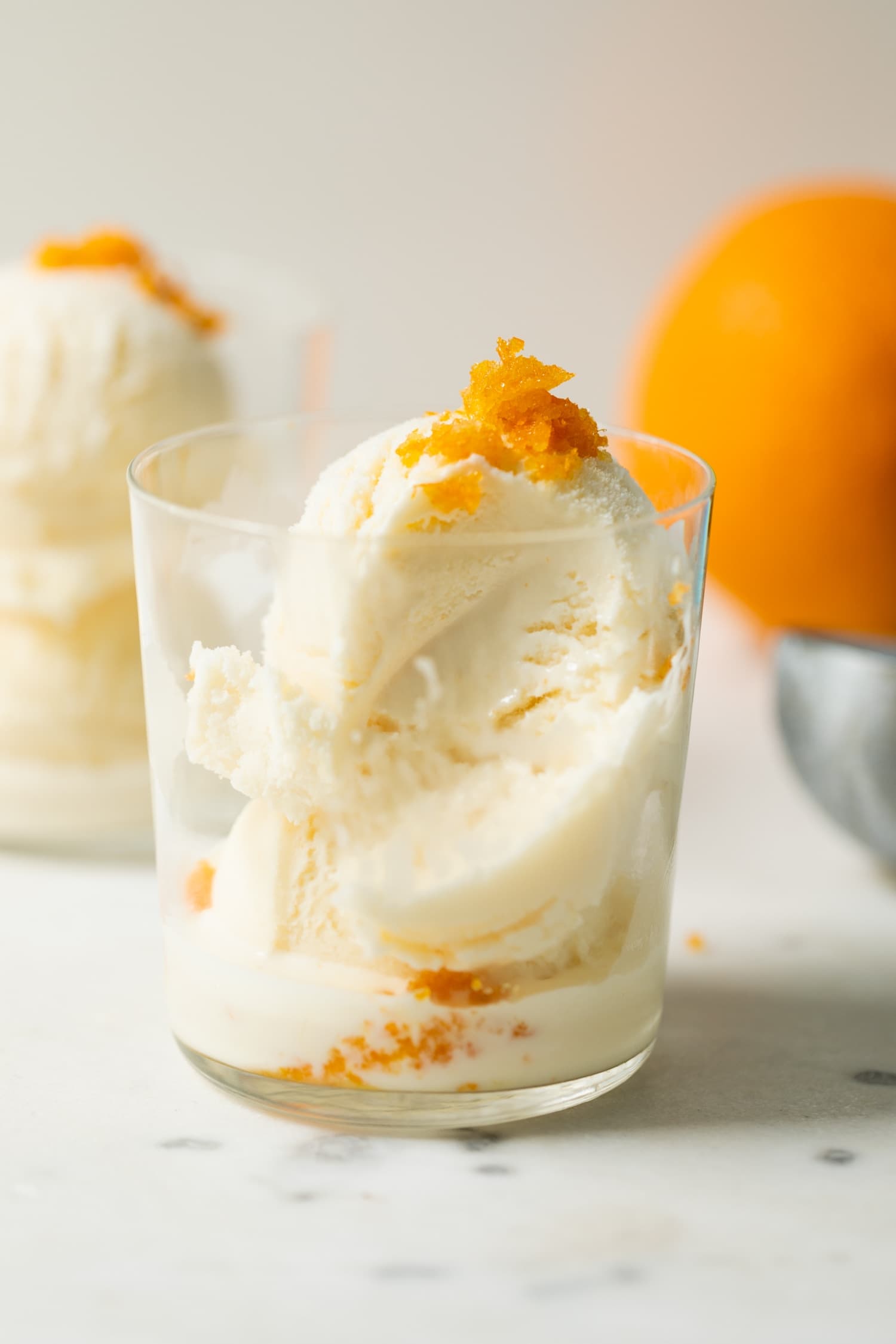 Mimosa ice cream speckled with orange zest pieces with a bite missing in a clear glass on marble surface with second ice cream glass and orange in background
