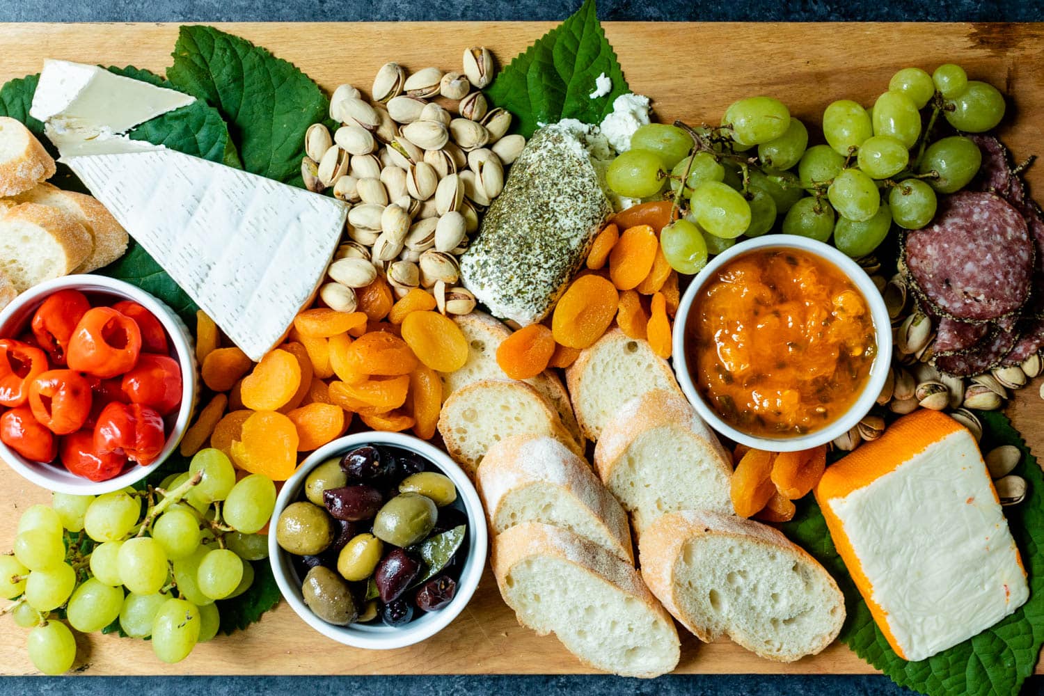 Top down view of peach jalapeno jam in a white bowl with a spoon surrounded by tomatoes, grapes, goat cheese, pistachios, olives, bread, dried apricots and meat all on a brown serving board with green leaves in the background