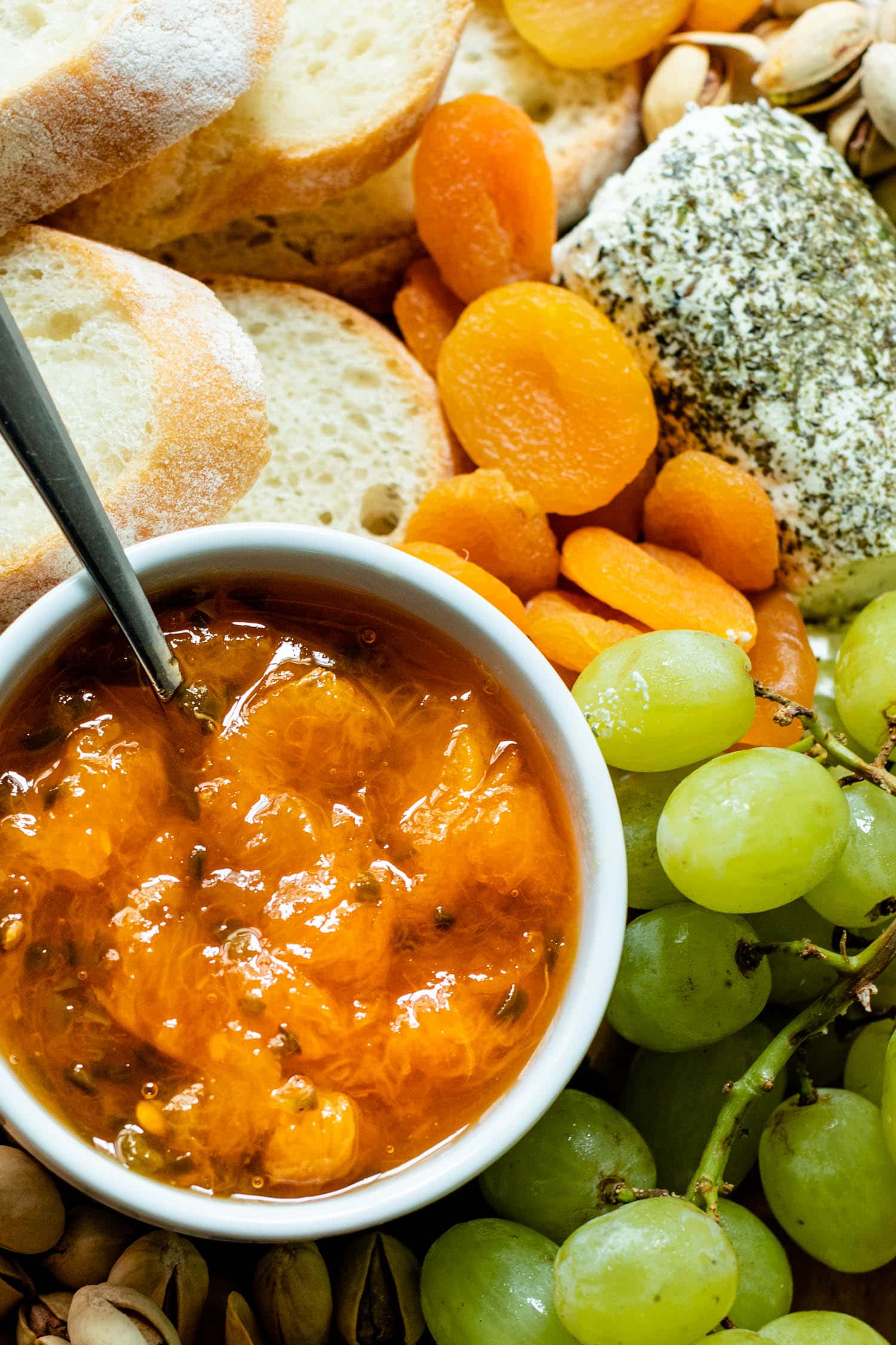 Top down view of peach jalapeño jam with a spoon in a white bowl surrounded by grapes, bread, dried apricots, cheese and goat cheese on a brown serving board