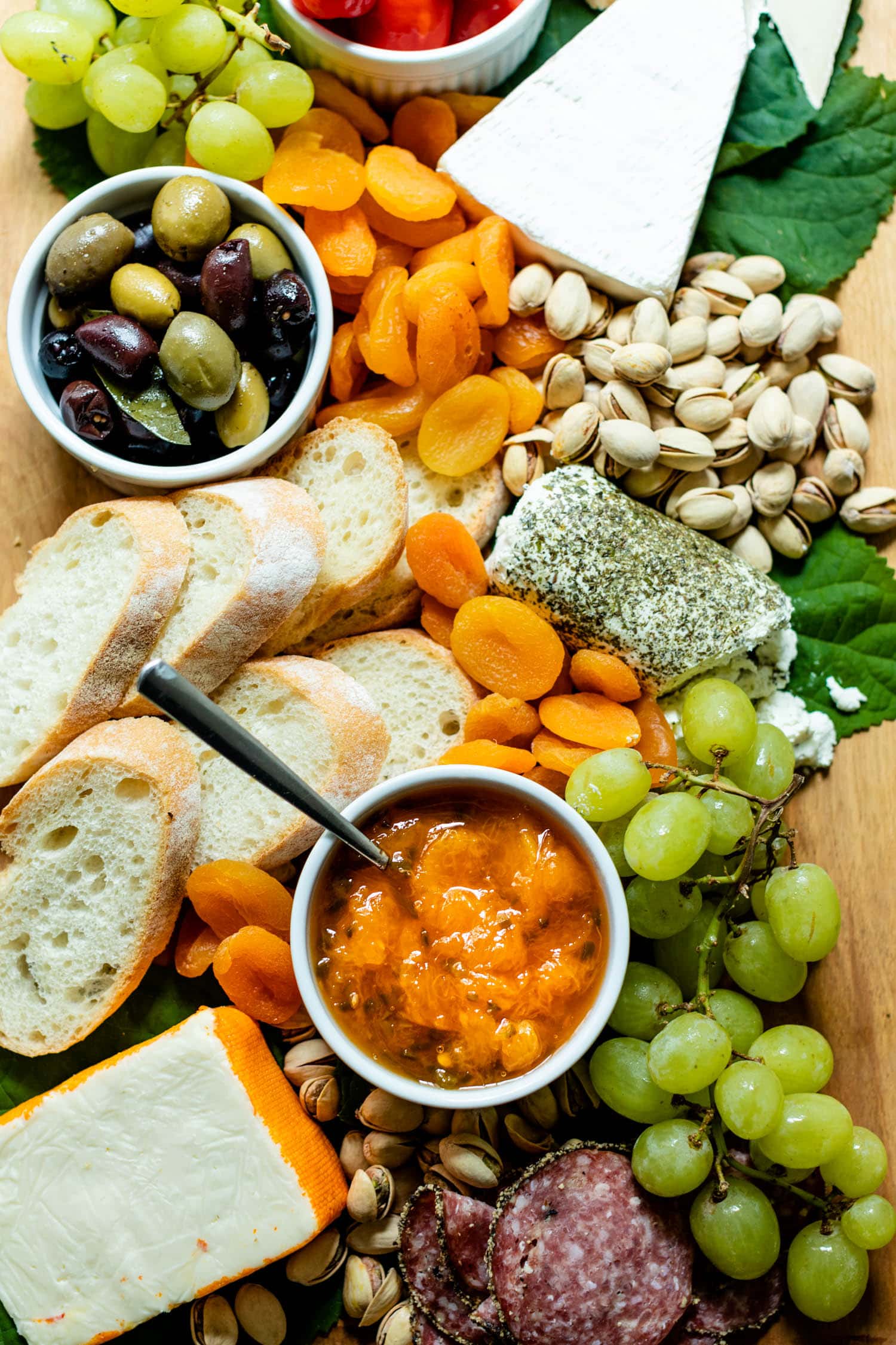 Top down view of peach jalapeño jam with a spoon in a white bowl surrounded by grapes, olives, pistachios, bread, dried apricots, cheese and goat cheese on a brown serving board