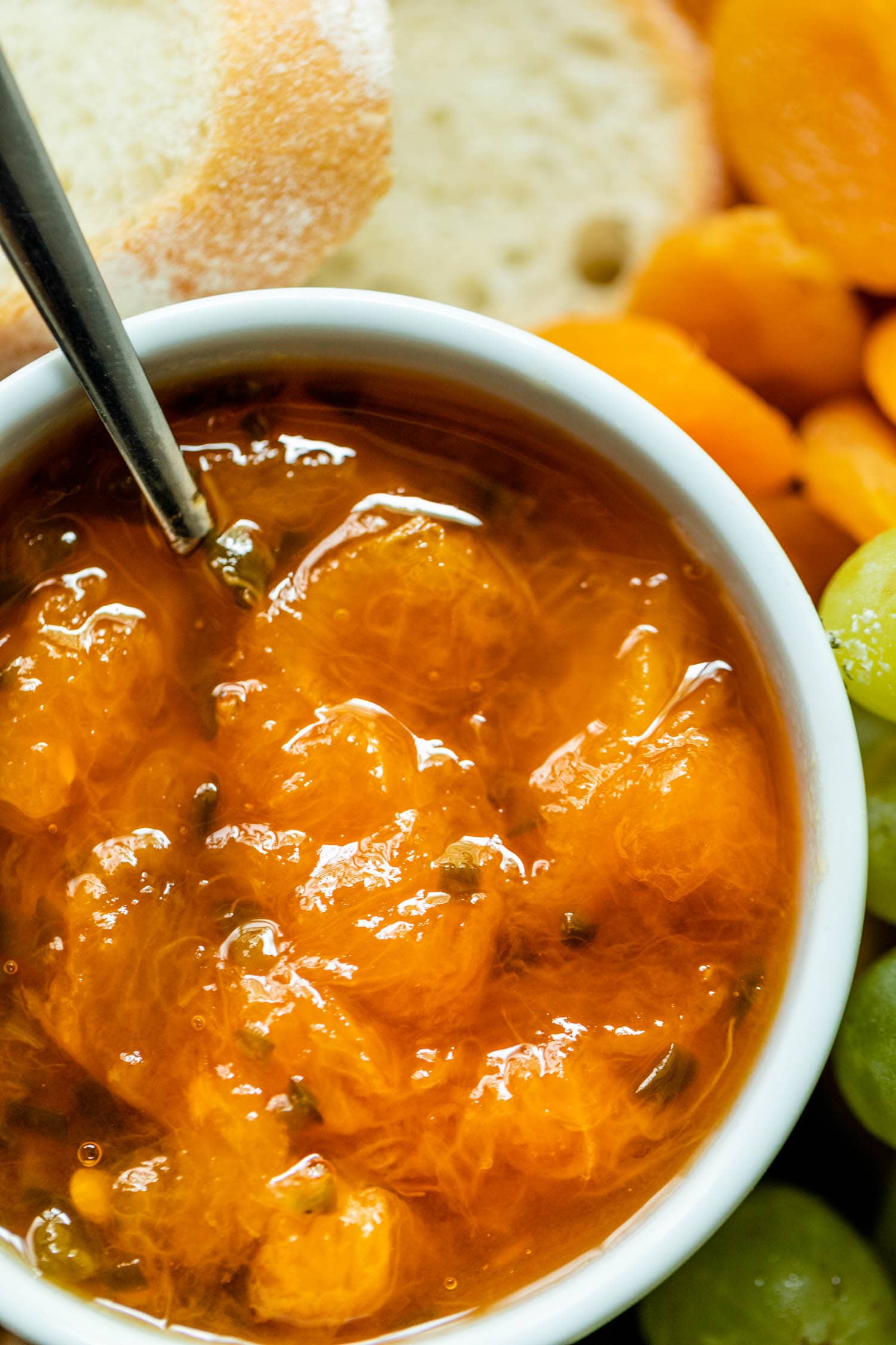 Close up view of peach jalapeño jam in a white bowl with a spoon surrounded by bread, grapes and dried apricots