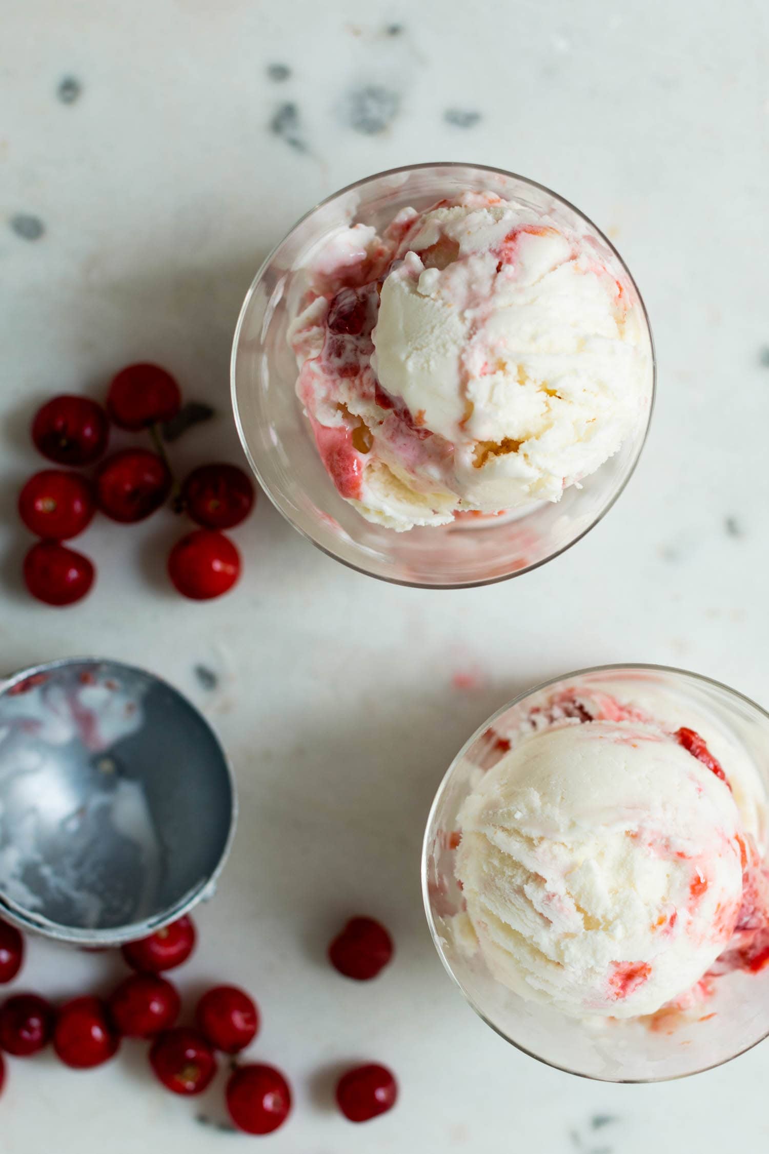 Top down view of two glasses filled with sour cherry frozen yogurt with streaks of cherry jam throughout with sour cherries and ice cream scoop in background on marble surface