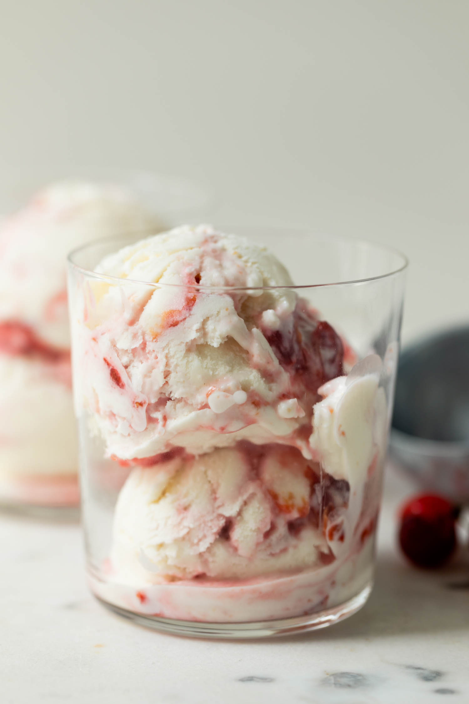 Sour cherry frozen yogurt with streaks of cherry jam throughout in clear glass with second glass, sour cherries and ice cream scoop in background on marble surface
