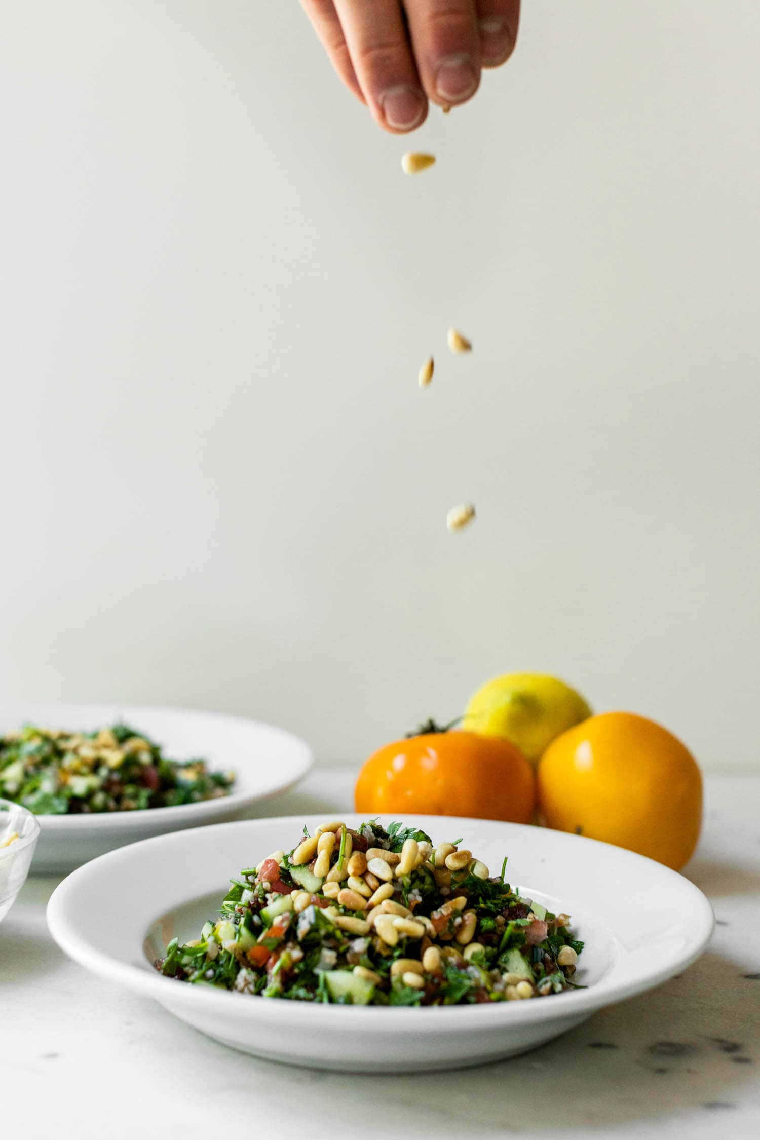 Bowl full of tabbouleh topped with pine nuts with hand at top of picture sprinkling more pine nuts on top with tomatoes and second bowl in background on marble surface
