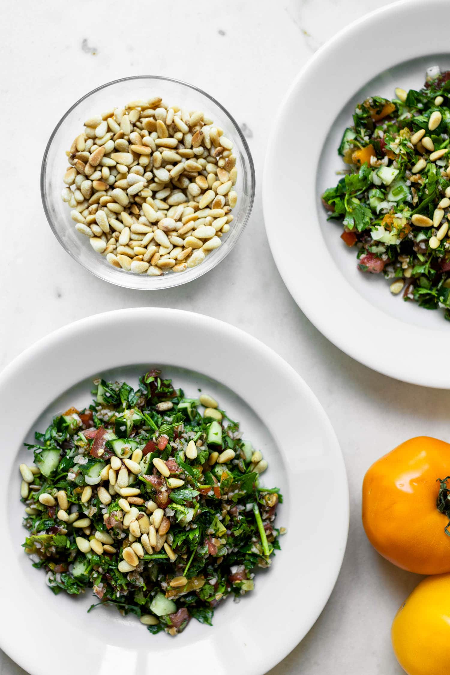 Top down view of two bowls full of tabbouleh topped with pine nuts with tomatoes and bowl of pine nuts on a marble surface