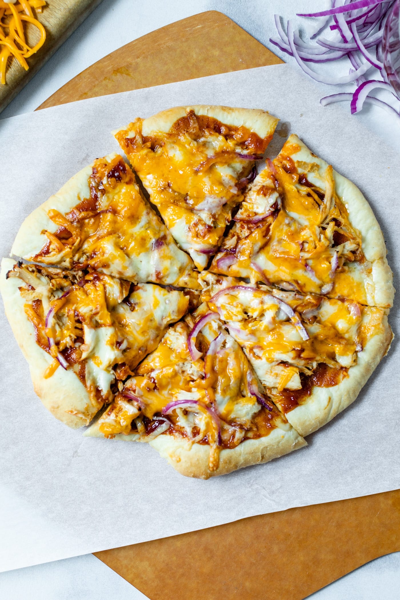 Top down view of sliced homemade barbecue chicken pizza made of chicken, cheese, barbecue sauce and red onion on a homemade crust on a piece of white parchment and pizza board with extra red onions and cheese on cutting board all on a marble surface