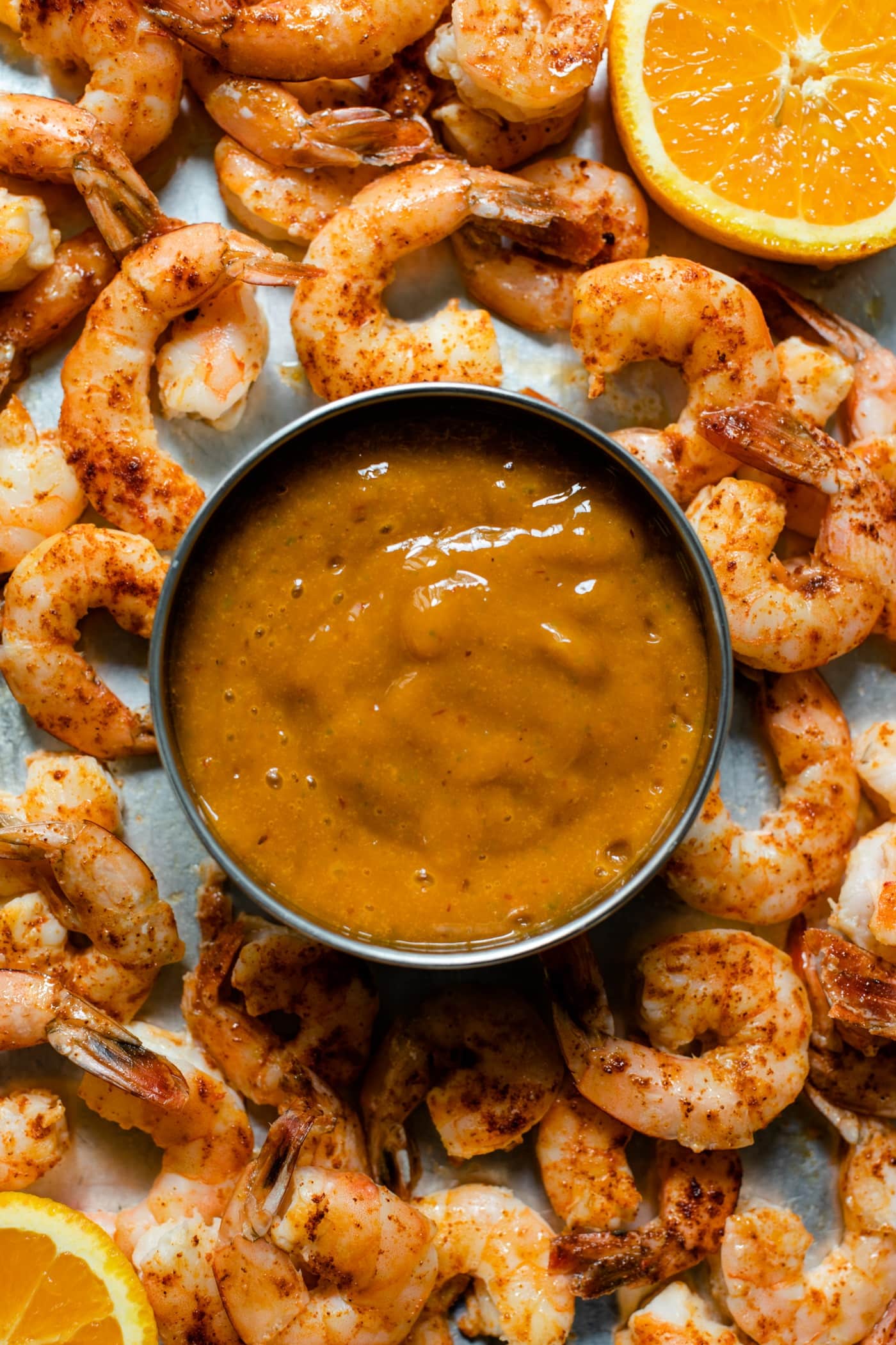 Top down view of pieces of chipotle shrimp on roasting sheet with slice of orange and bowl of mango sauce