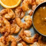 Top down view of pieces of chipotle shrimp on roasting sheet with slice of orange and bowl of mango sauce on a gray concrete surface