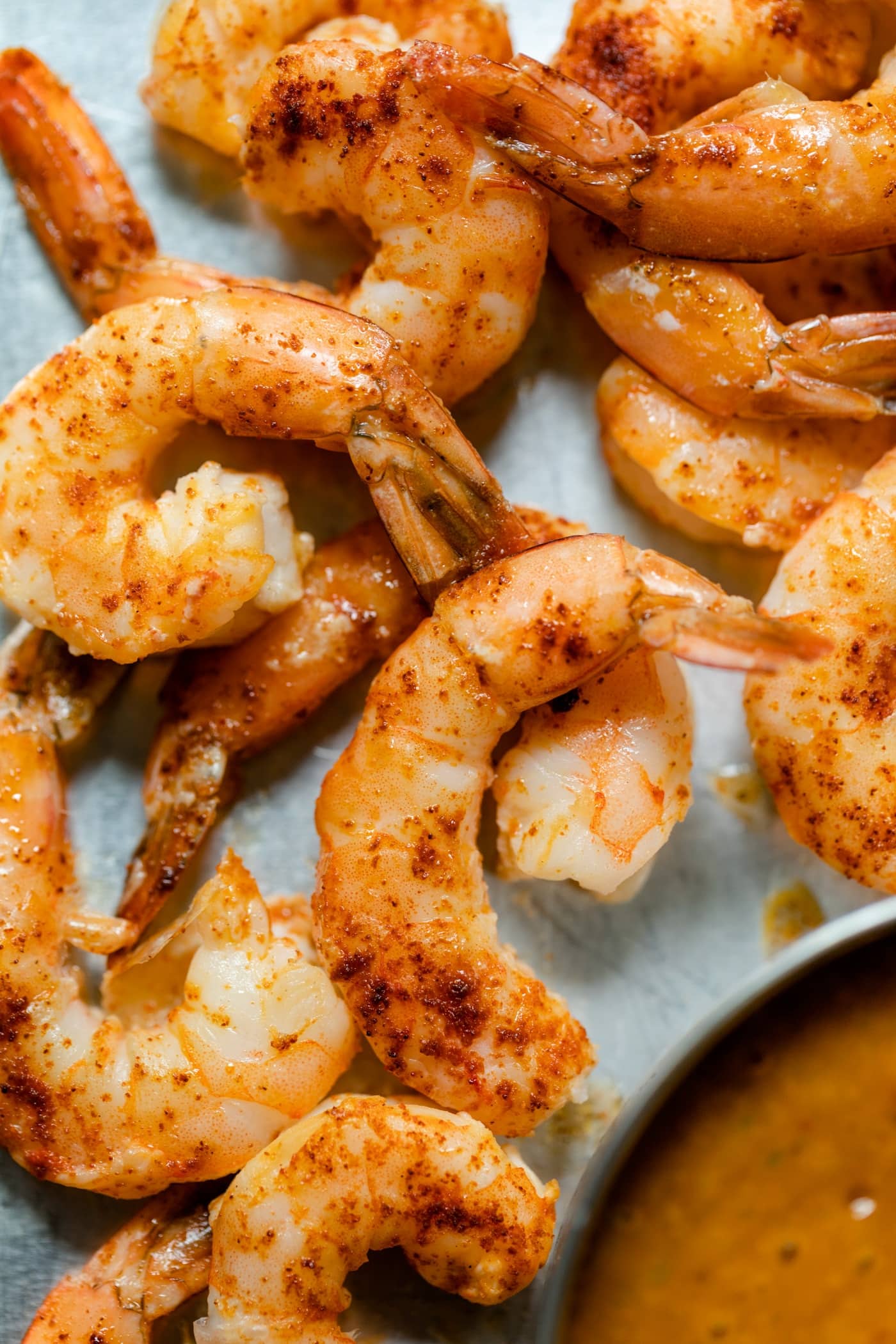 Top down view of pieces of chipotle shrimp with tails attached on roasting sheet with bowl of mango sauce on a gray concrete surface