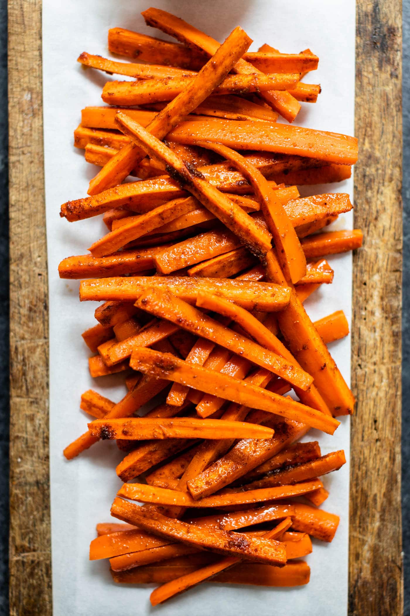 Orange harissa roasted carrots piled on a piece of parchment on a brown cutting board all on gray concrete surface