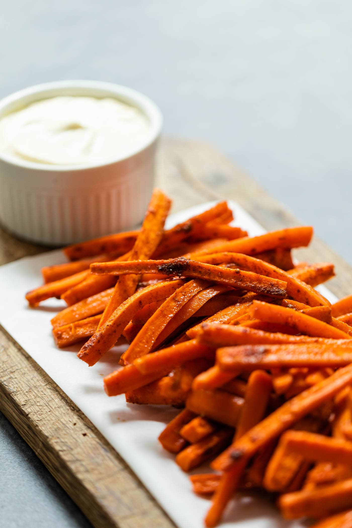 Orange roasted carrots piled on a piece of parchment on a brown cutting board with bowl of garlic lemon aioli all on gray concrete surface
