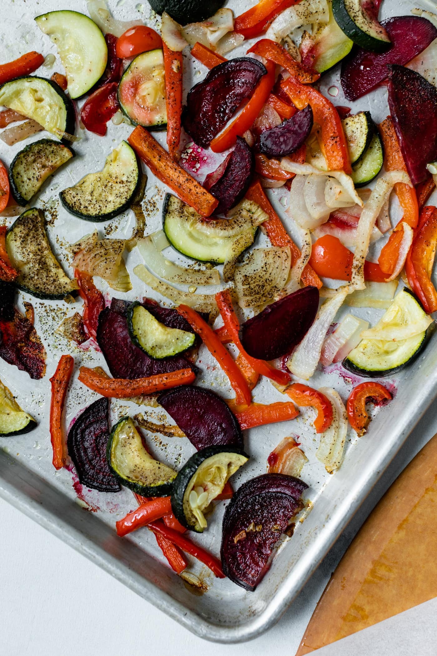 Top down view of roasted vegetables on sheet pan for homemade roasted vegetable pizza with pizza board and piece of parchment on marble surface