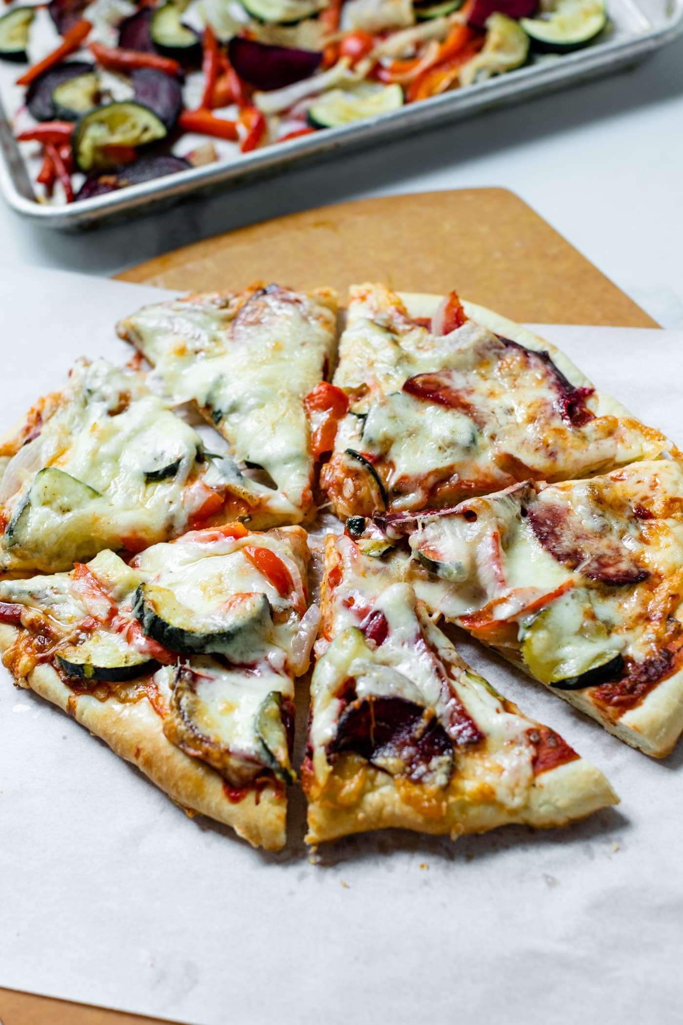 Pieces of homemade roasted vegetable pizza in circle on top of piece of white parchment and pizza board with pan of roasted vegetables in background all on marble surface