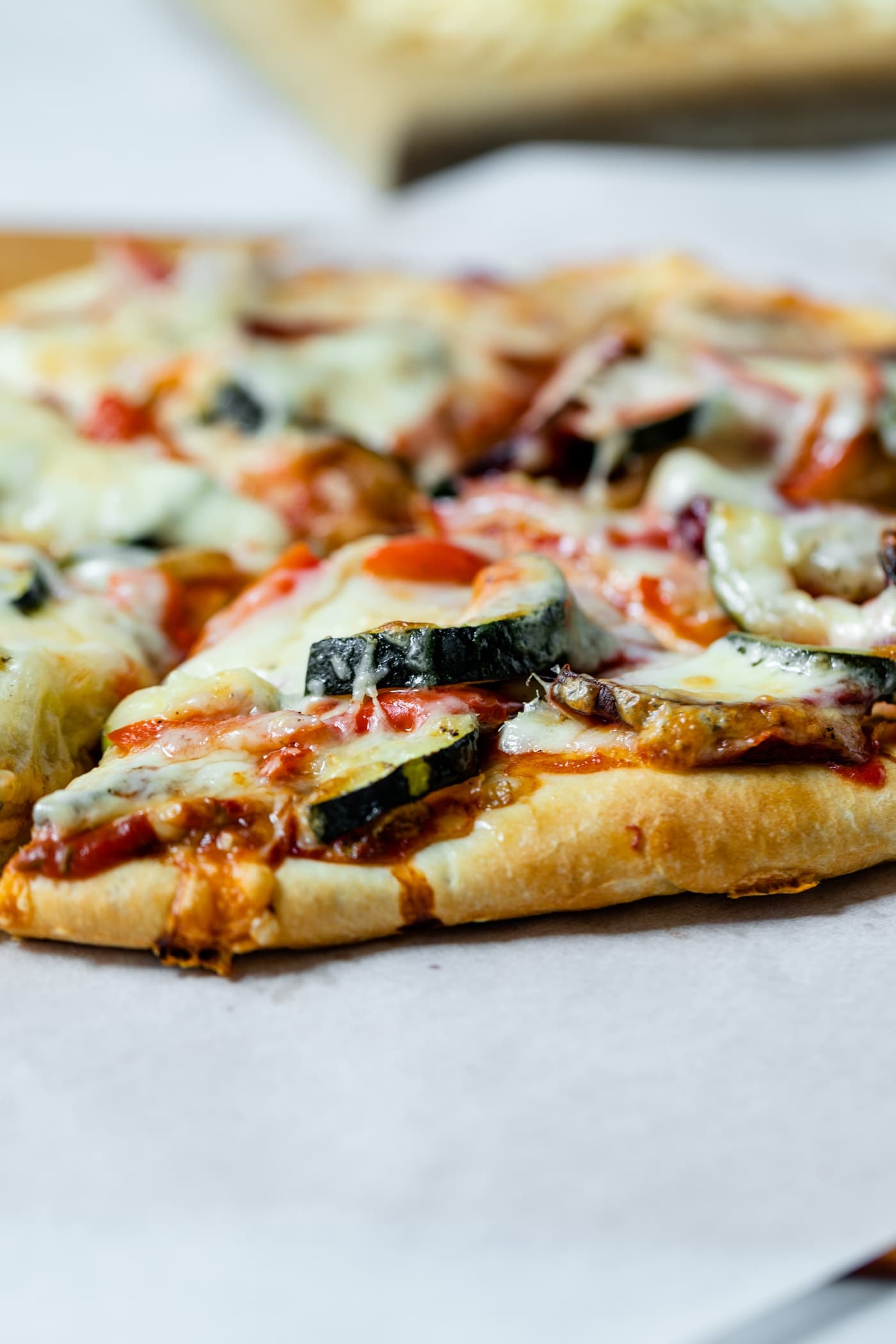 Homemade roasted vegetable pizza with vegetables and cheese on crust on top of piece of white parchment with cutting board and cheese in background