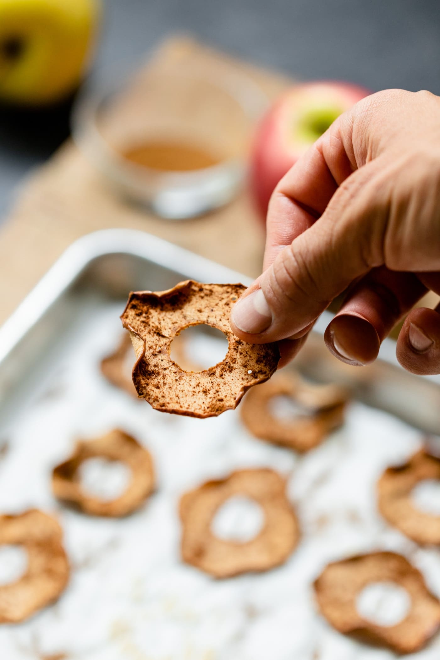 Hand holding homemade apple chip with sprinkling of spices on top with additional apple chips in background on a wood board on a gray surface