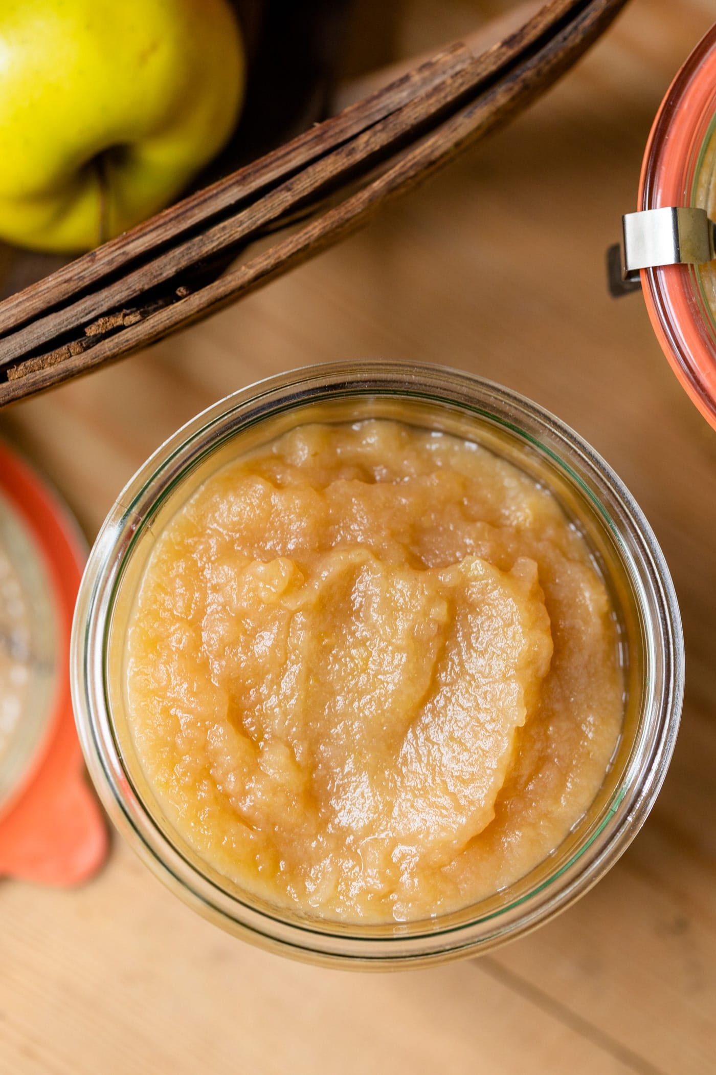Top down view of glass jar filled with homemade applesauce on wood board with basket filled with apples and extra glass jars