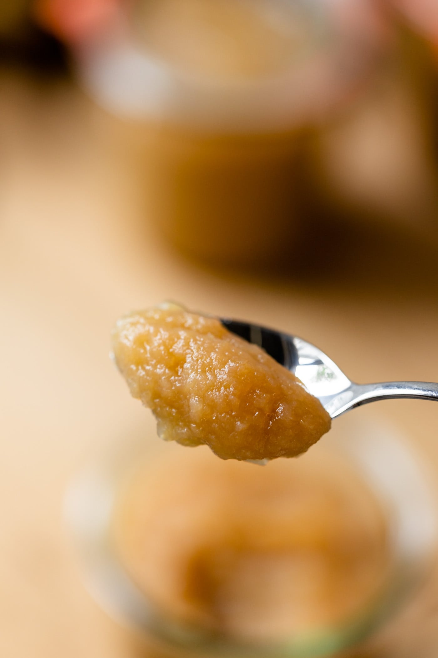 Homemade applesauce sitting on silver spoon with glass jar filled with applesauce in background on wood board