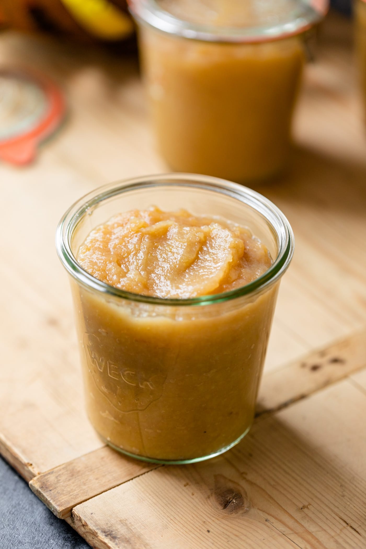 Glass jar of homemade applesauce sitting on wood board glass jar in background on gray slate surface