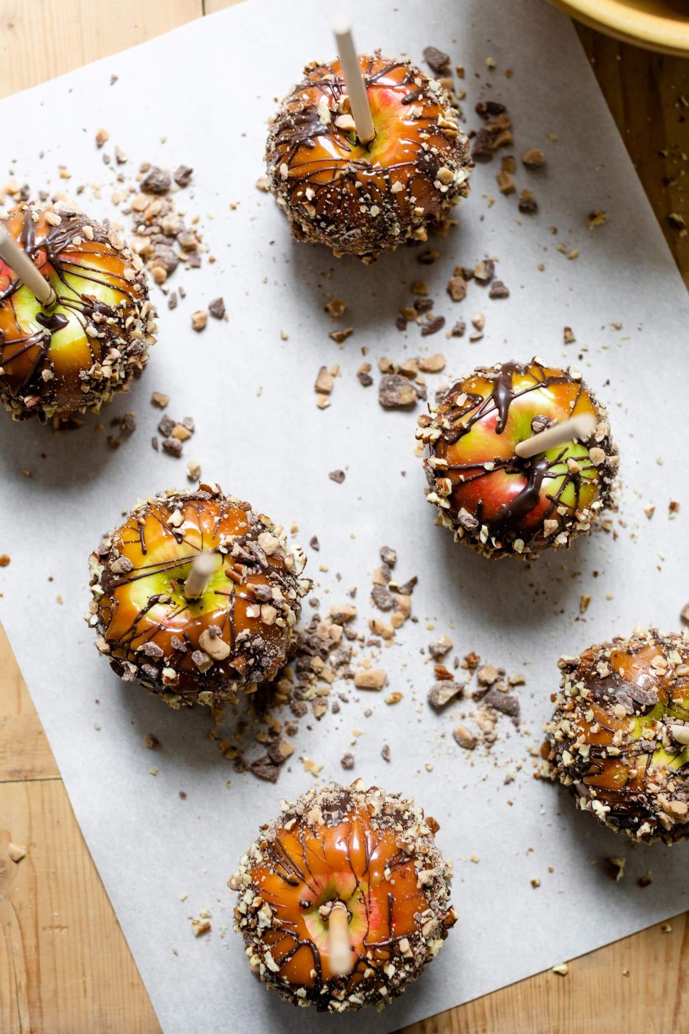 Homemade caramel apples sitting on a piece of white parchment sprinkled with toffee pieces and chopped pecans all on a wood board