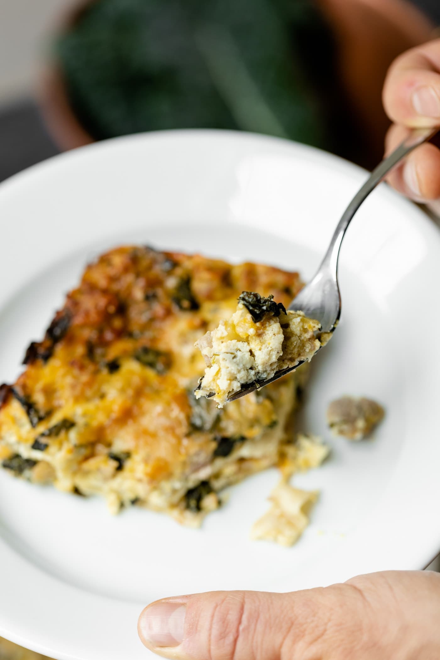 Piece of squash kale lasagna held by hand on a white plate with fork holding piece of dish with copper pot and kale in background on gray slate surface