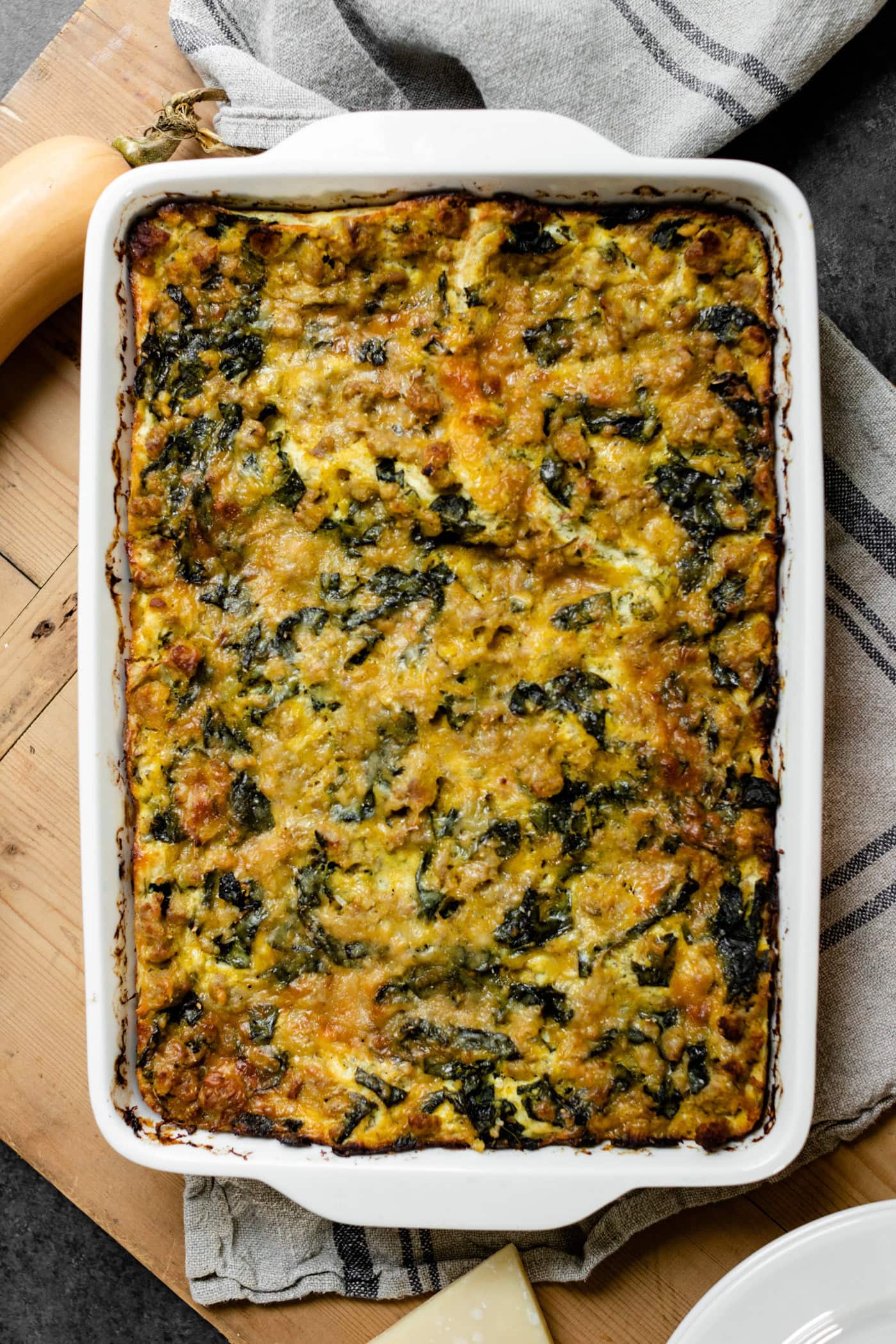Top down view of squash kale lasagna in white baking dish sitting on wood board with Parmesan cheese, squash and towel underneath all on gray slate surface