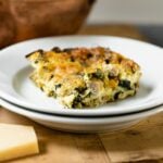 Piece of squash kale lasagna sitting on white plate with extra white plate underneath with piece of Parmesan cheese on wood board with copper pot in background all on gray slate surface