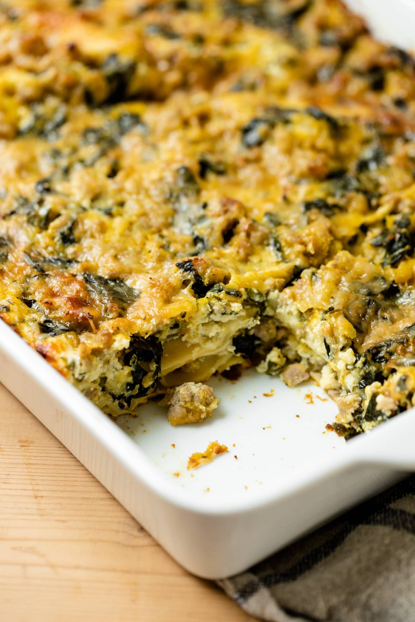 Squash kale lasagna sitting in white baking dish with one piece removed on a wood board with linen towel underneath