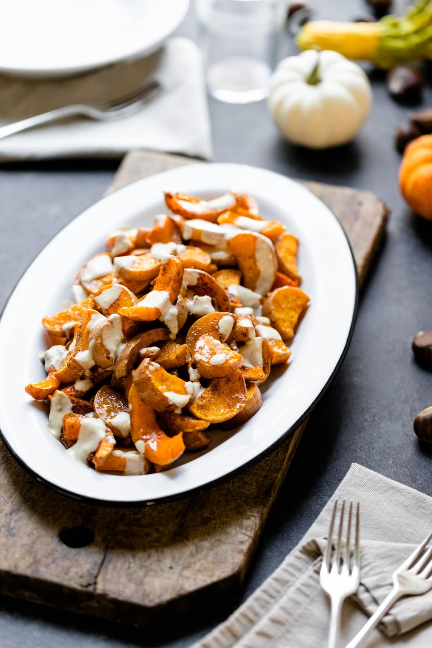 Spiced butternut squash drizzled with white tahini sauce on white platter sitting on wood board with extra plates, glasses and silverware on gray slate surface