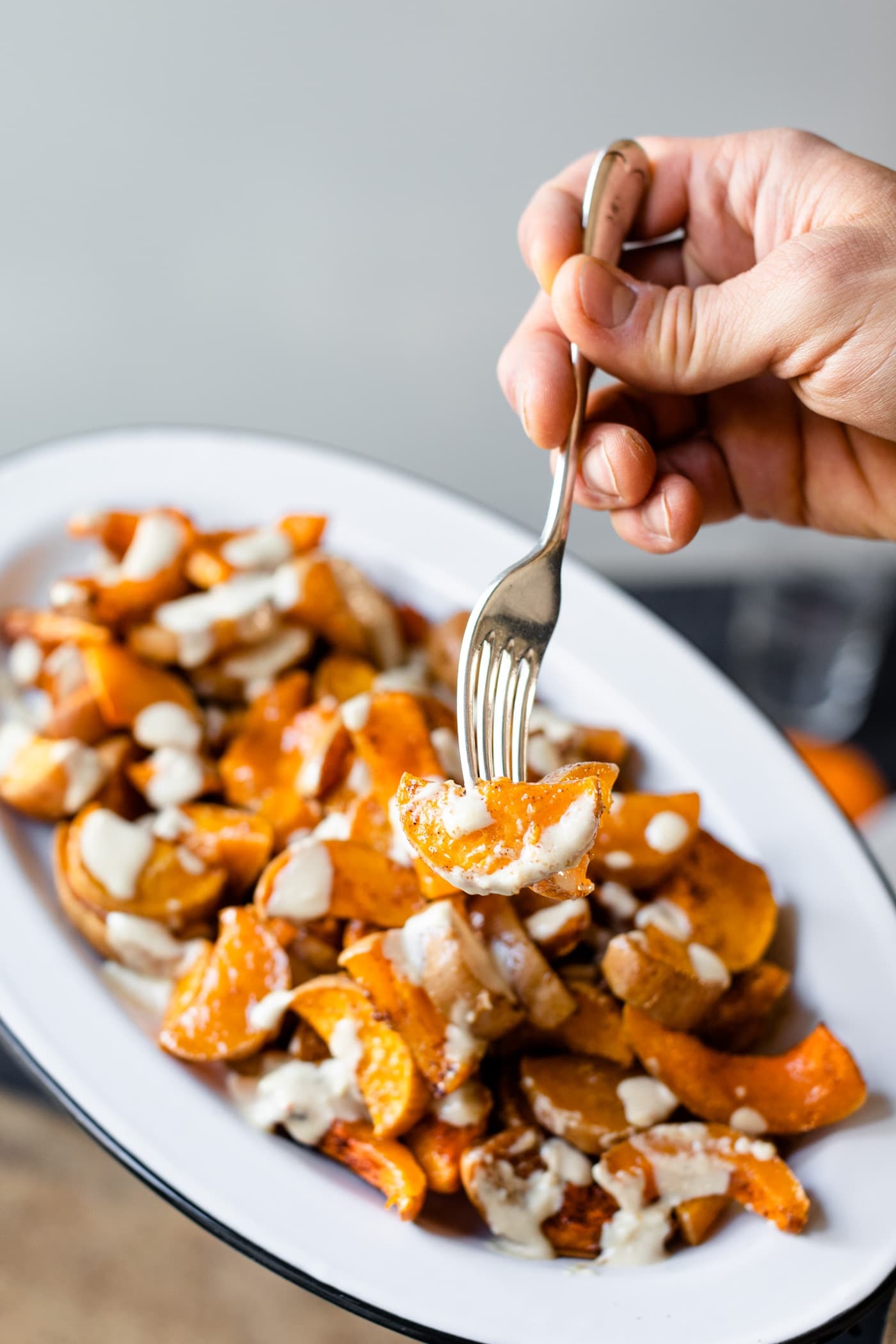 Piece of spiced butternut squash drizzled with white tahini sauce on a fork being held with platter in background on gray slate surface