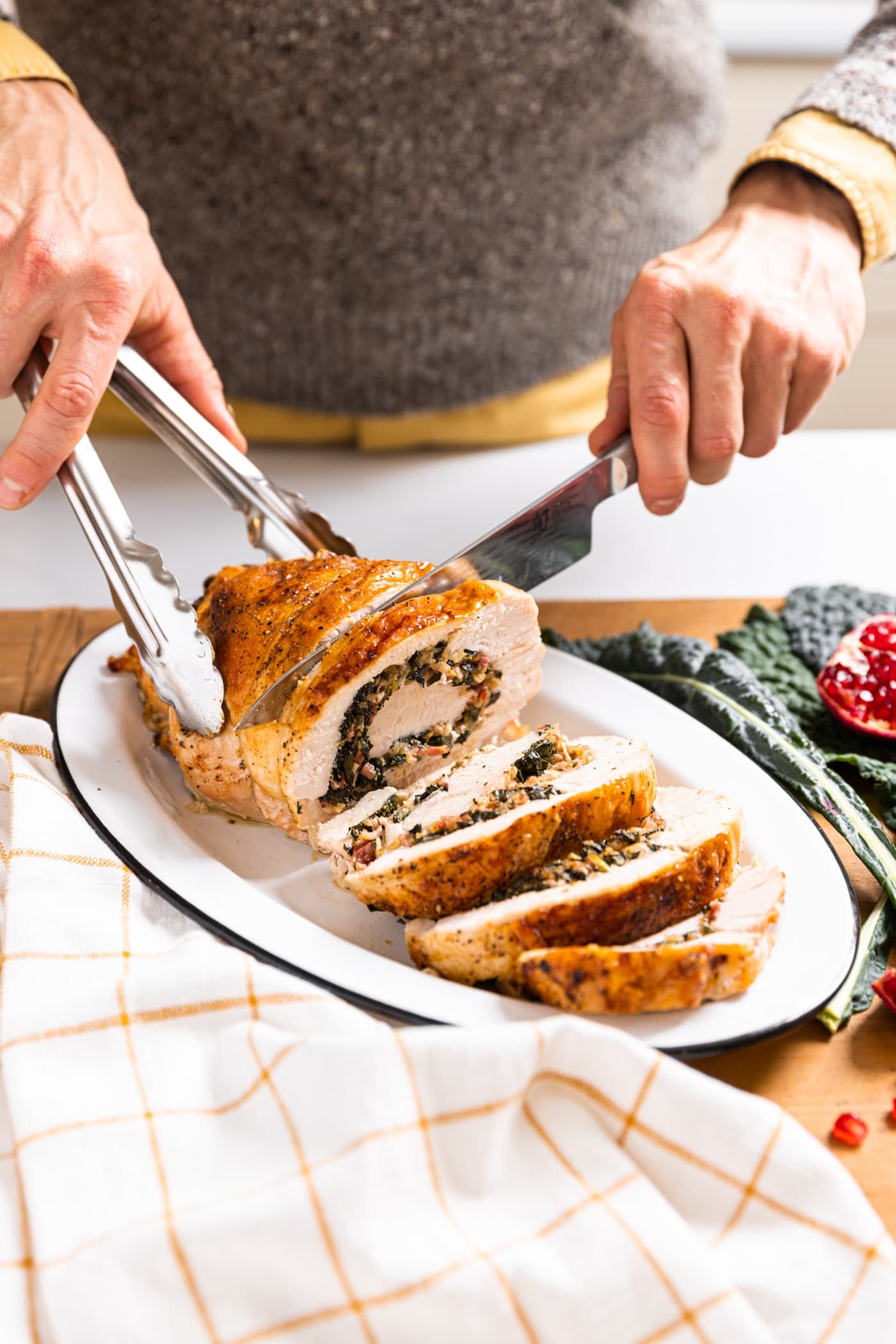 Two hands slicing slices of turkey roulade with green colored kale stuffing onto white plate on white countertop