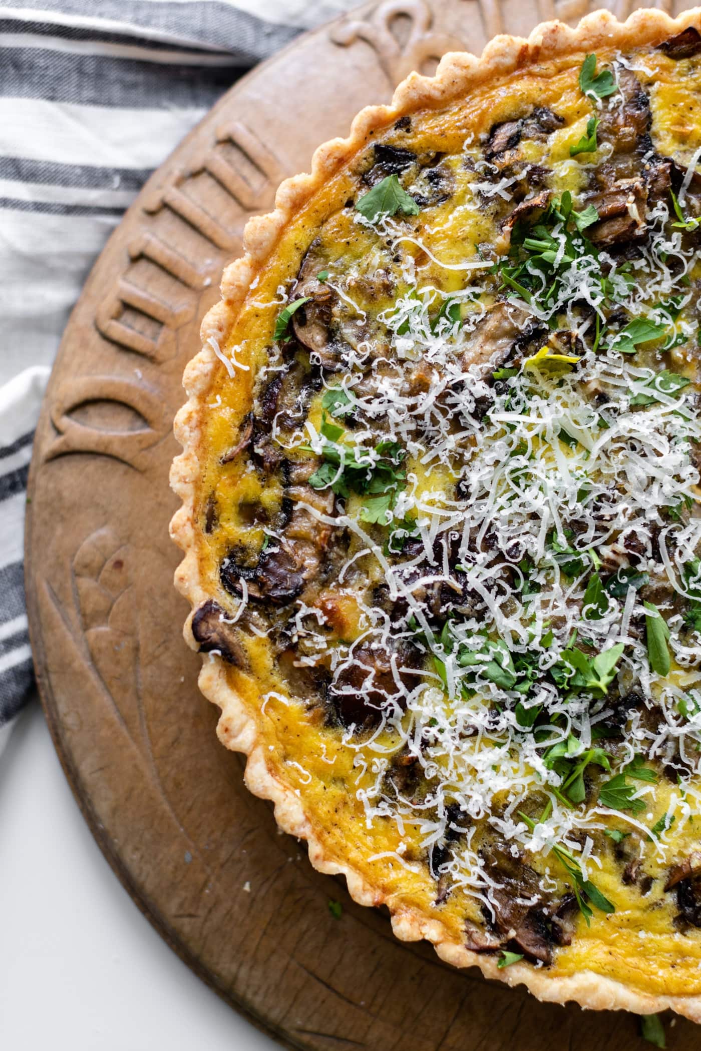 Breakfast quiche topped with parsley and Parmesan cheese sitting on bread board with linen underneath all on white surface