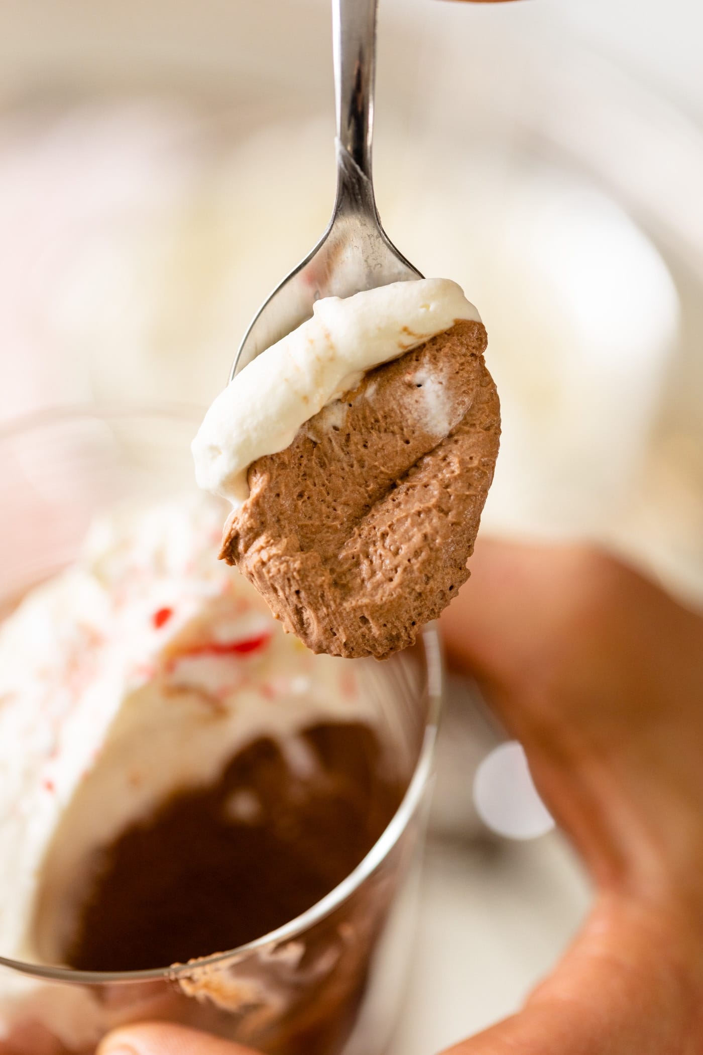 Chocolate mousse sitting on spoon held by hand with extra glasses of chocolate mousse topped with whipped cream and peppermint bits in background.