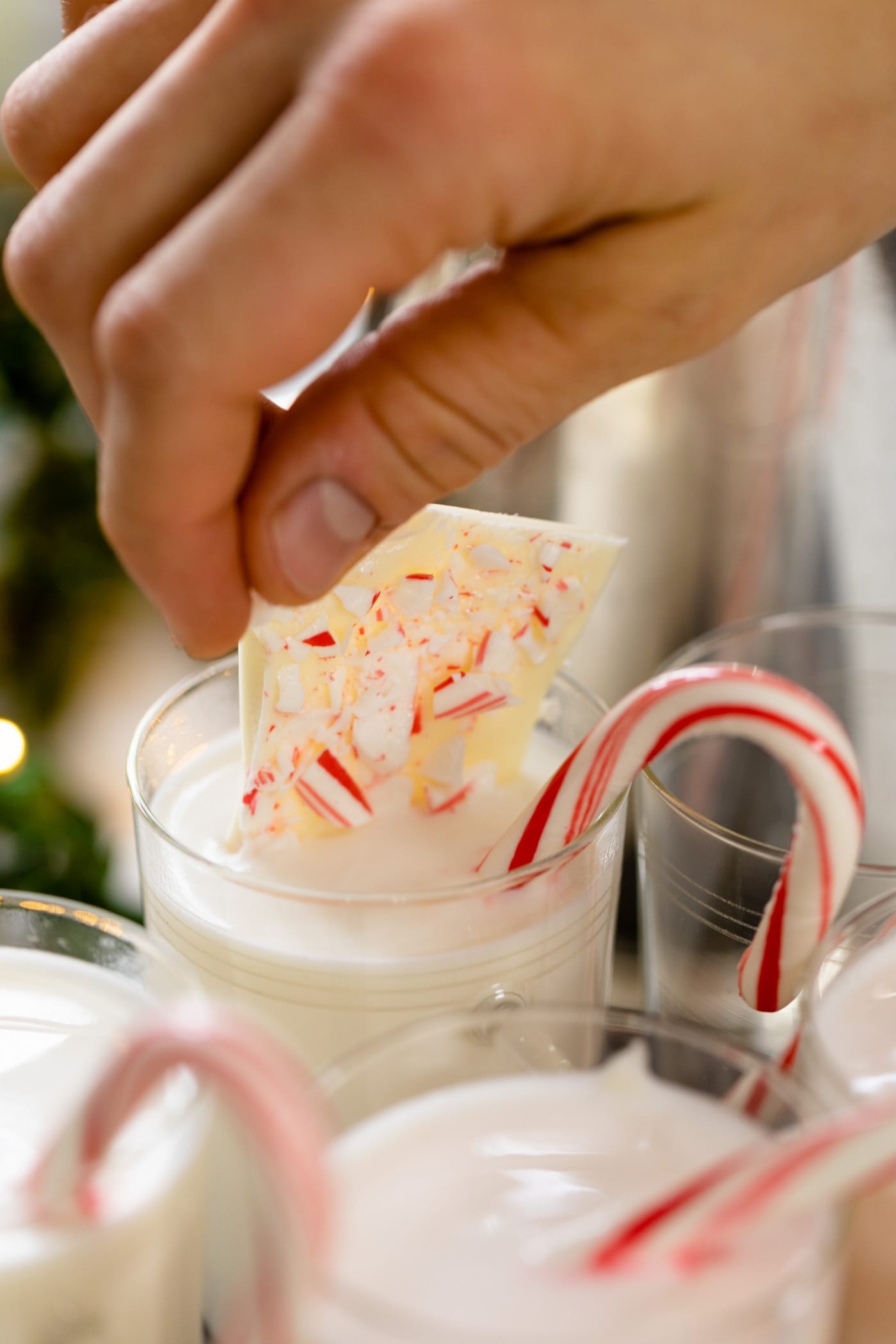 Hand putting piece of peppermint bark into glass full of white hot chocolate with candy cane sitting on edge of glass
